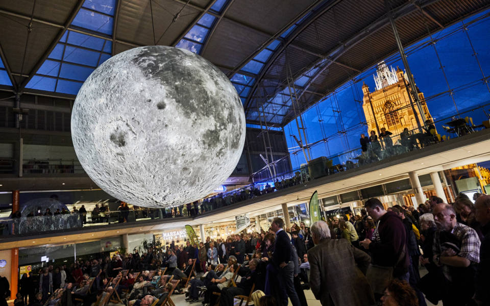 Picture taken inside The Forum, in Norwich. St Peter Mancroft Church can be seen, illuminated, in the background. A giant inflatable moon hangs in the middle of The Forum. People are underneath the moon, looking up at it.