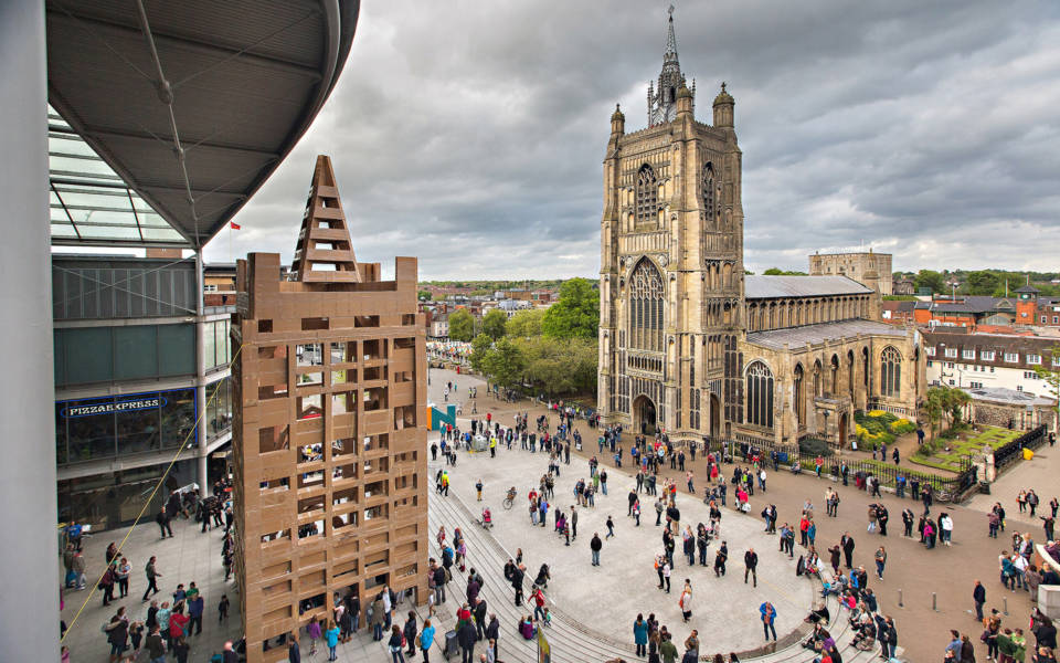 Picture of Millennium Plain in Norwich. Photo is taken from a tall building, looking down on the Plain. St Peter Mancroft Church is on the right side of the picture. A large replica of the church, made from cardboard boxes is on the left side of the picture. Over 100 people are walking around the Plain.