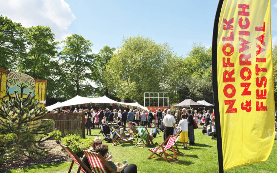 Picture taken in the Adnams Spiegeltent compound. People sit in deckchairs and other people are queuing to get into the Spiegeltent. A large yellow and red NNF flag is in the foreground of the image.