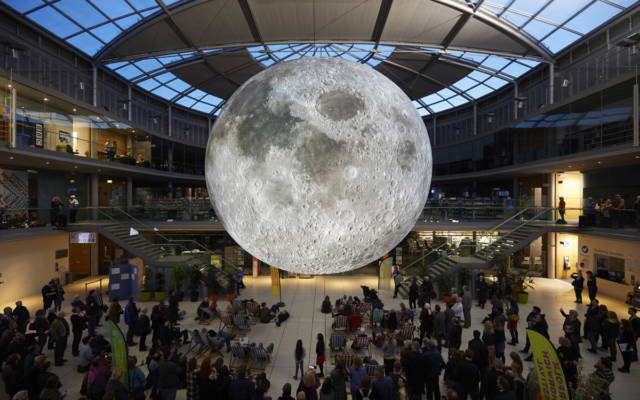 Picture inside the Forum, Norwich. A giant inflatable moon hangs in the middle of the building, it is illuminated. Approx. 100 people stand beneath it, some people sit in deckchairs.