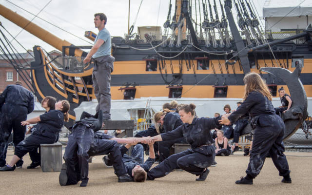 A group of dancers in blue boiler suits perform in front of a ship