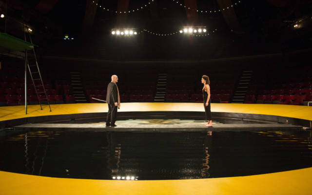 Production photo from NNF16 show The Tempest, two people stand facing each other. Raked seating surrounds them on all sides.