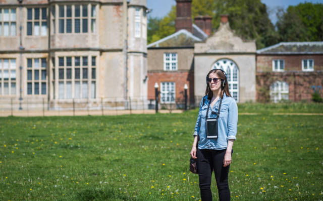 Photo from NNF16 show Walk With Me, a woman stands in front of Felbrigg Hall, she is wearing headphones and an ipad hangs around her neck.
