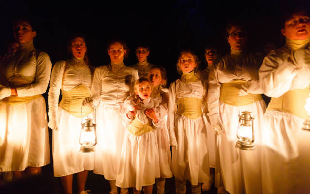 Photo from NNF15 show Wolf's Child, a group of women and young girls stand in a line. They are wearing white dresses and corsets, four of them are holding lanterns, it is night time.