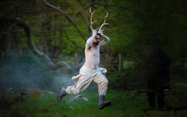 Photo from NNF15 show Wolf's Child, a man painted in white, holds two antlers to his head and runs through a woodland area.