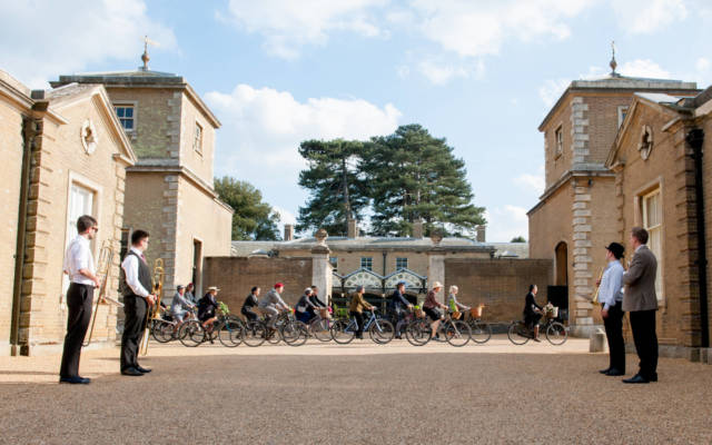 Photo from NNF14 show Souvenir, a group of approx 10 people ride bikes through the courtyard at Holkham Hall.