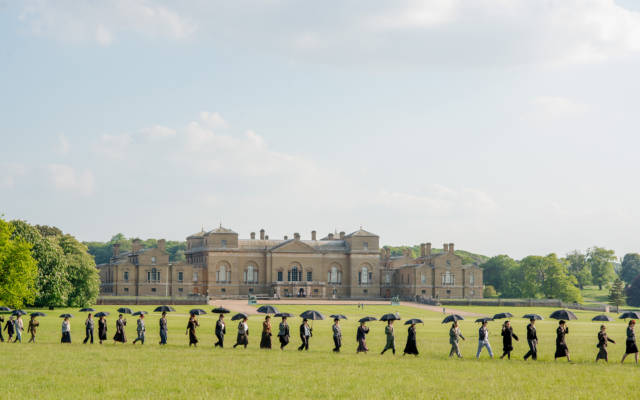 Photo from NNF14 show Souvenir, a long procession of people walk through a field, each holding a black umbrella. Holkham Hall is visible in the background.