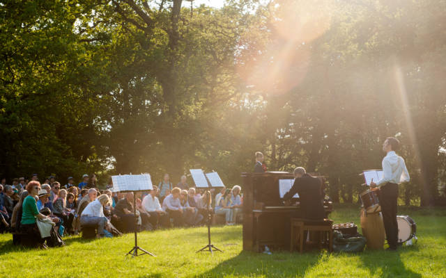 Photo from NNF14 show Souvenir, set outside in a wooded area on a sunny day. A large crowd watch a man playing a piano.