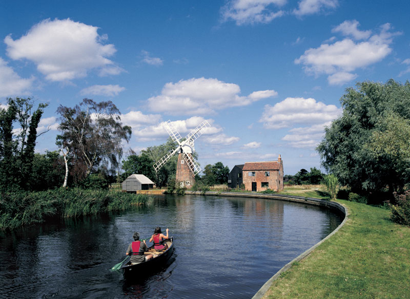 Two people canoe down a bending river, it is a sunny day. A windmill can be seen in the distance.
