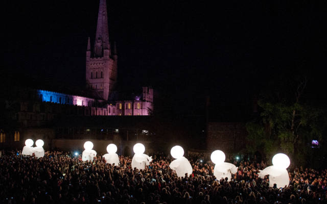 Photo from NNF13 show Reve dHerbert. It is night time. In the background there is Norwich Cathedral, which is illuminated in a purple/blue light. In the foreground there are hundreds of people, all gazing at eight large, white, glowing, inflatable figures.