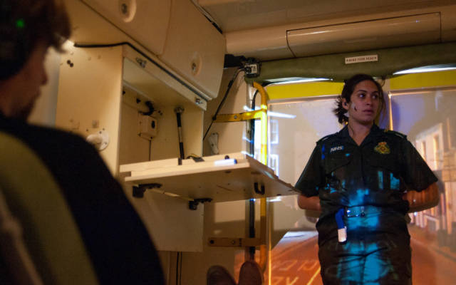 Photo from NNF13 show The Kindness of Strangers, a man wearing headphones sitting in the back of an ambulance. A paramedic stands with her back to the doors of the ambulance, an image of a street is projected over her.