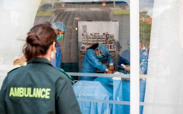 Photo from NNF13 show The Kindness of Strangers, a paramedic looks through a window. Inside the building there are four people performing surgery.