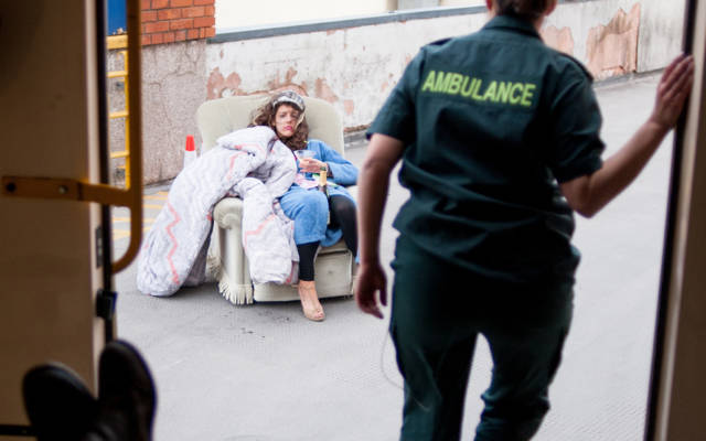 Photo from NNF13 show The Kindness of Strangers, taken from inside an ambulance with the back doors open, a paramedic steps out of the ambulance. There is a woman sitting on a chair on the pavement outside.