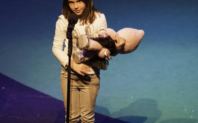 Photo from NNF12 show 100% Norfolk, a little girl stands on stage holding a pink toy. She is talking into a microphone.