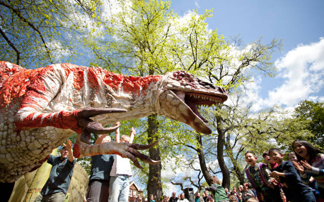Photo from NNF12 show Dinosaur Petting Zoo, a huge T-rex puppet faces an audience of children who are all standing up and clapping.
