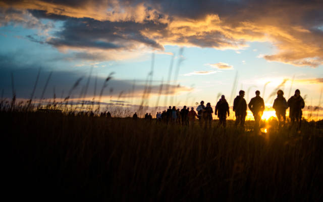 Photo from NNF13 show Ideas of Flight, a line of people walk in pairs, the sun is setting behind them.