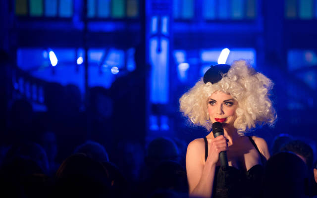 Photo from NNF13 show Les Enfants Perdus, a woman wearing a big blonde wig and a tiny black beret holds a microphone to her mouth.