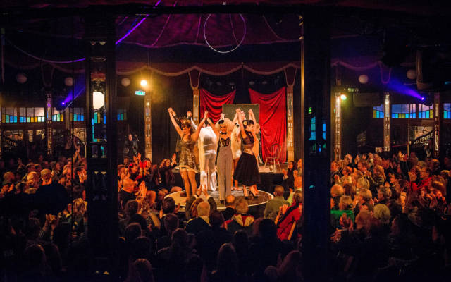 Photo from NNF13 show Les Enfants Perdus, a group of performers stand on a circular stage in the spiegeltent, a busy crowd surrounds them, clapping.