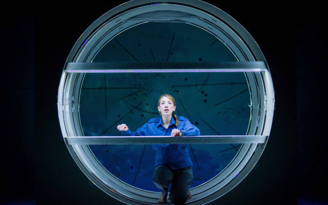Photo from NNF14 show Pioneer, a woman dressed in blue with her hair in a plait kneels in a big circular space, staring at the camera.