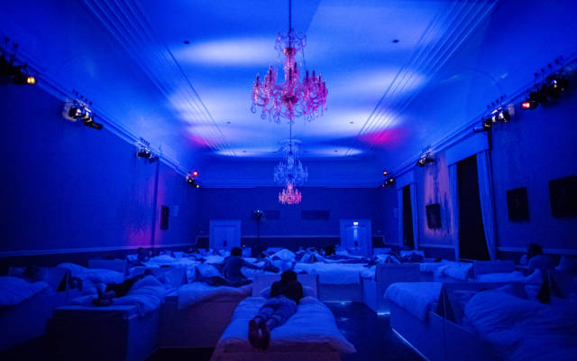 Photo from NNF17 show The Arms of Sleep, a room filled with beds is illuminated in blue light. Chandeliers hang from the ceiling.