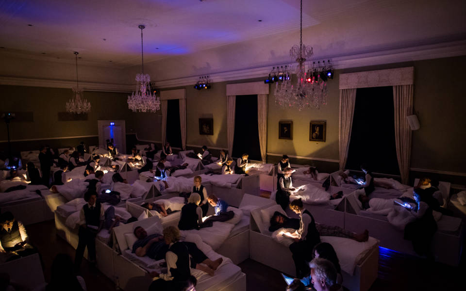 Photo from NNF17 show The Arms of Sleep, approx. 40 beds are arranged in a large room. Audience members lie one per bed, and members of the choir sit on the end of each bed.