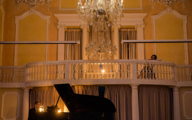 Photo from NNF17 show The Arms of Sleep, taken inside a grand room with yellow walls and a chandelier hanging from the ceiling. A man is playing piano on stage.