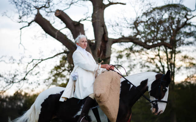 A woman dressed in white rides a black and white horse, a large tree with only a few leave is behind her.