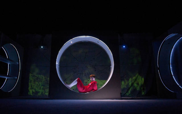 A woman in a red boiler suit sits in a large block on stage. The background is a projection of a woodland area.