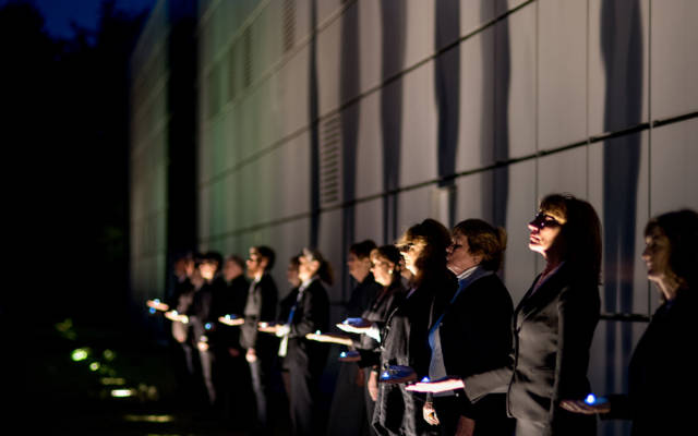 The Voice Project Choir lined up outside the Sainsbury Centre for Visual Arts, they are each holding a blue light in one hand and are illuminated from spotlights in the ground.