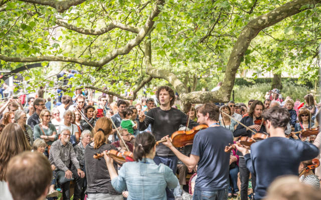 An orchestra perform outside, under a large tree. Lots of people are watching them. It is a sunny day.