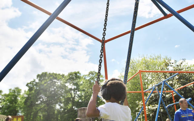 A boy sits on a swing, wearing a VR headset. Lots of people are visible in the background of the image.