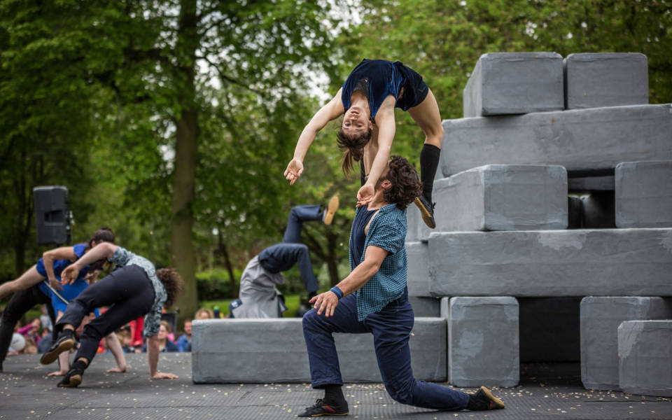 Image from an NNF16 show. Lots of large grey blocks are stacked on top of each other on the right side of the picture. A woman is held up by a man who is kneeling in the foreground of the image.