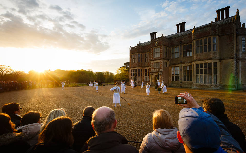 Image from Wolf's Child, a 2015 Festival show. Audience members stand in a semi-circle, watching women wearing long white dresses. Felbrigg Hall is in the right side of the picture.