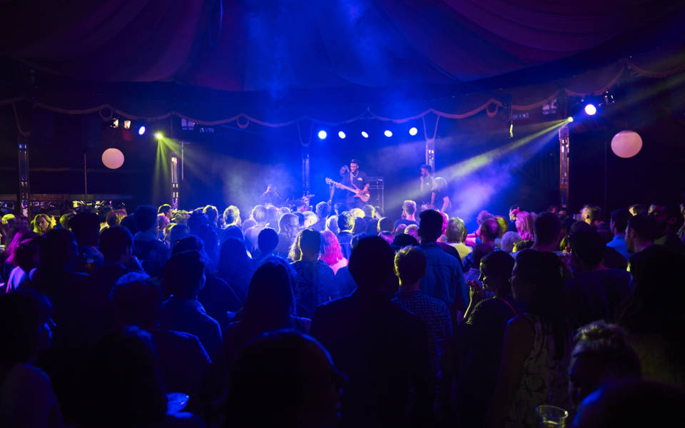 Picture taken inside the Adnams Spiegeltent. A five piece jazz band 'Ezra Collective' are on stage, a busy crowd, illuminated in blue light, is watching them.