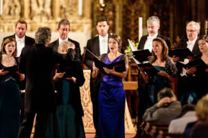 The Sixteen choir stand in a row, they are all singing.