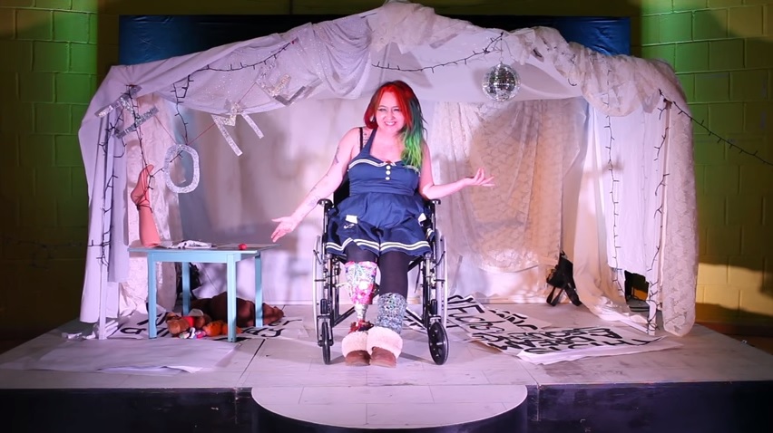 A woman sits in a wheelchair, she is on stage with a white tent behind her.