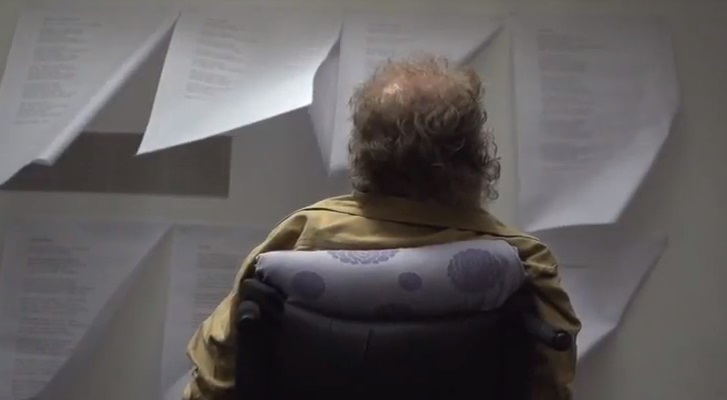 A man sits with his back to the camera, he is looking at some sheets of paper that are tacked to the wall.