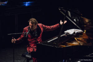 Chilly Gonzales sits at a piano in a red smoking jacket and ascot, holding a microphone he gestures emphatically at the crowd