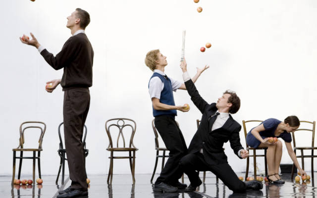 Two men stand back to back juggling apples, whilst a women sits down holding apples and man strikes a pose with a newspaper.