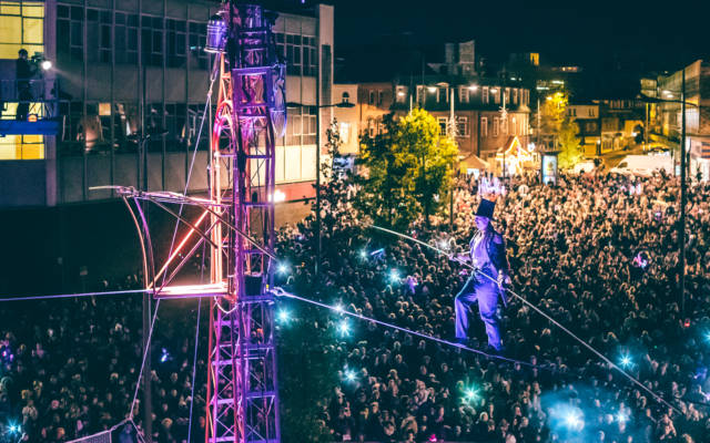 Chris Bullzini tight-rope walking at night, above a big crowd, illuminated by purple lights