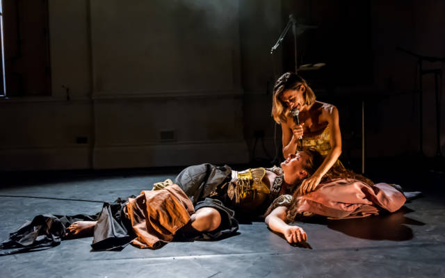 Photo: dressed. production shot, one woman sitting on the floor, holding a microphone, another lying on the floor, head resting on first woman's lap