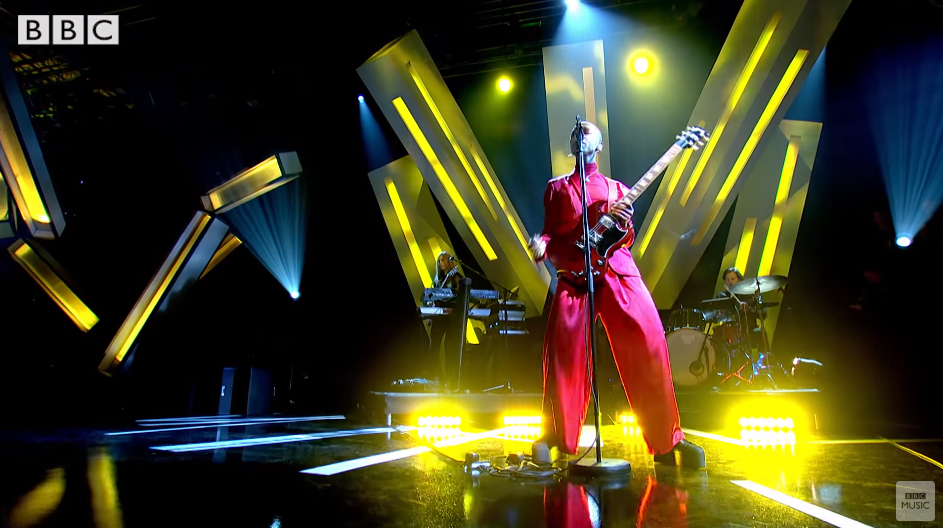 Video thumbnail: Nakhane onstage wearing a red suit with baggy trousers and plateau shoes playing the guitar. Drummer and someone playing the keyboard in the background. Stage is illuminated by yellow lighting
