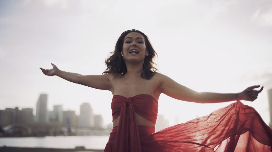 Video thumbnail: Nella wearing a flowy red dress, arms stretched out and singing to the camera. Cityskape in the background