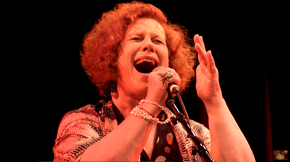 Photo: Sarah Jane Morris singing into a microphone, illuminated by warm orange lighting