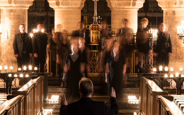 Photo: Tenerae choir in a church, conductor has his back to the camera. Choir is slighlty blurred in a ghostly manner.