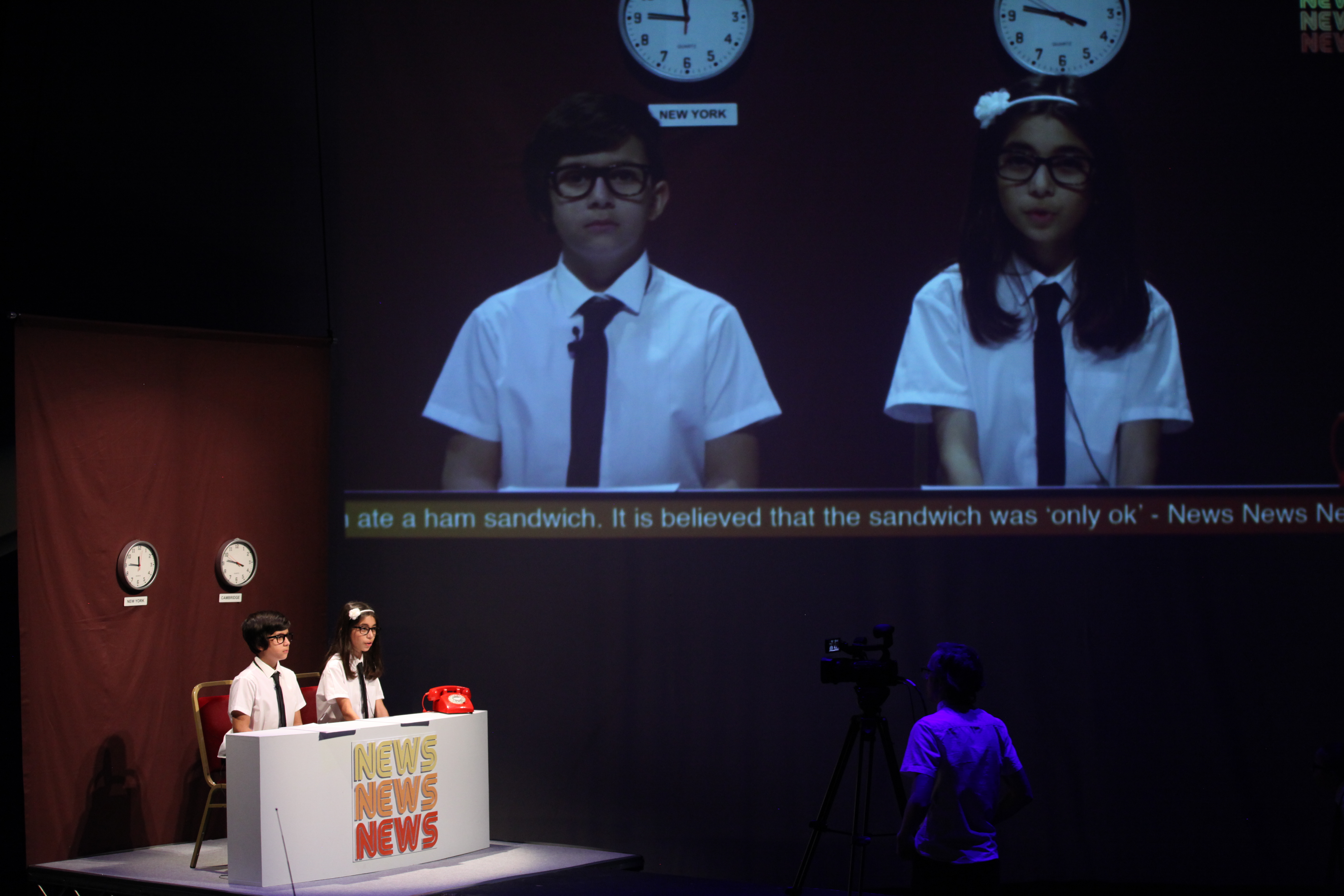 A boy and a girl sitting at a news desk on stage in the lefthand corner. They're shown on a big screen in the middle of the stage.