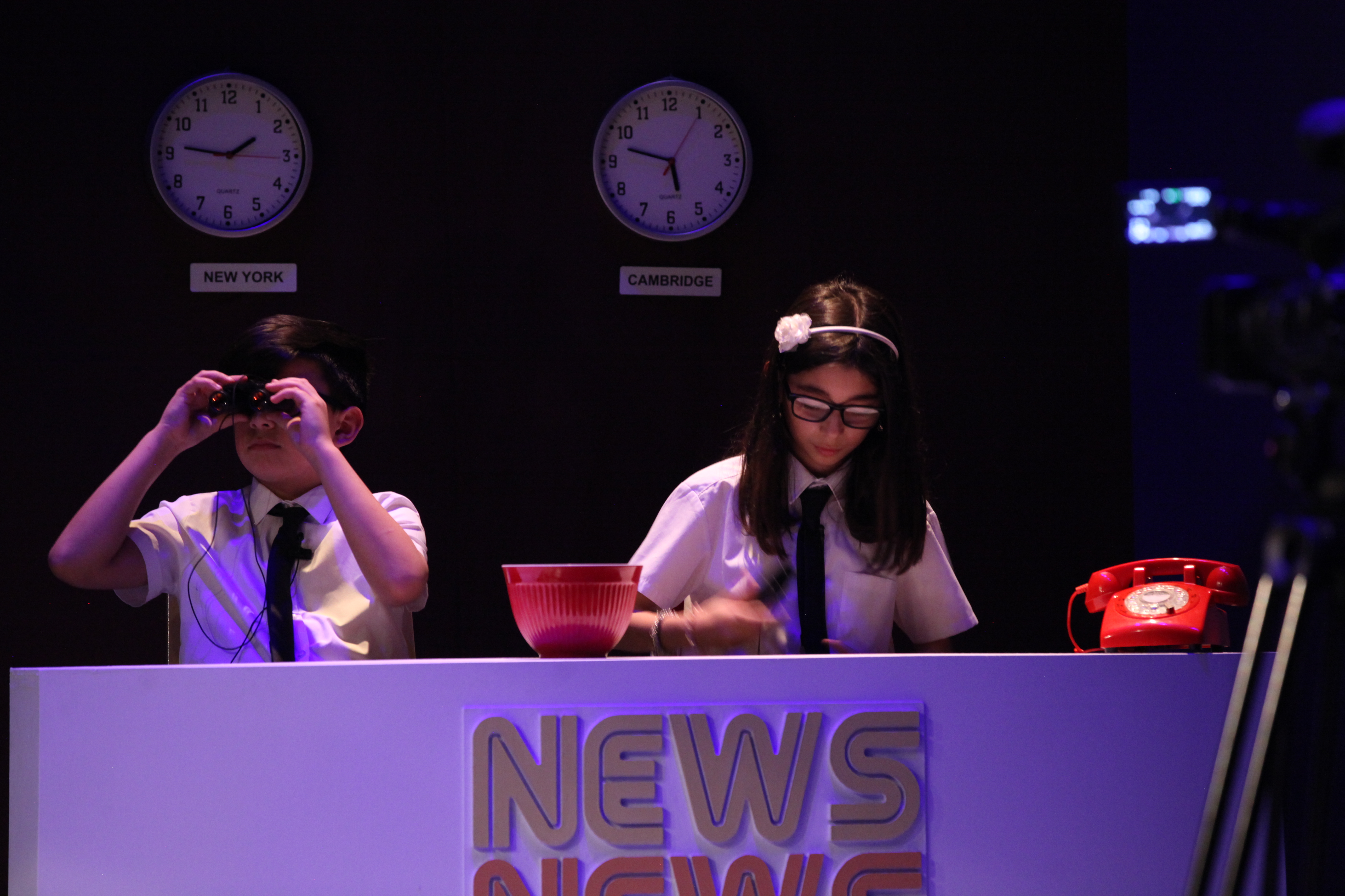 Two children wearing white shirts and black ties, sitting at a news desk