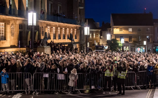 A large crowd standing behind barriers, in front of Norwich City Hall, looking to the sky to see the highwire walk.
