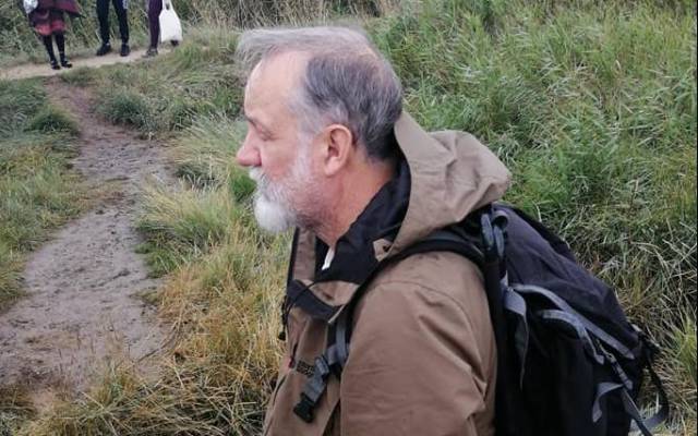 Ian Brownlie stands in overgrown grass, wearing rucksack and looking to left of the image