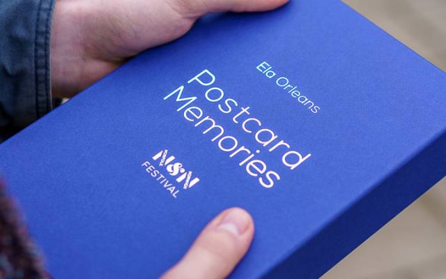 Hands hold a blue box with 'Postcard Memories' in silver text